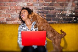 young hipster african American woman works at home, sitting on couch using laptop while her pet puppy dog looks on.