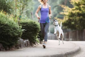 A middle aged women enjoys a morning run on a beautiful sunny day with her pet, the dog appreciating the time outside. They jog through a suburban neighborhood, the dog misbehaving and biting the leash.